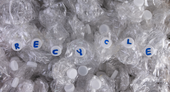 The word 'RECYCLE' is spelled out using bottle caps on a pile of crushed plastic bottles, promoting the recycling of plastic waste.