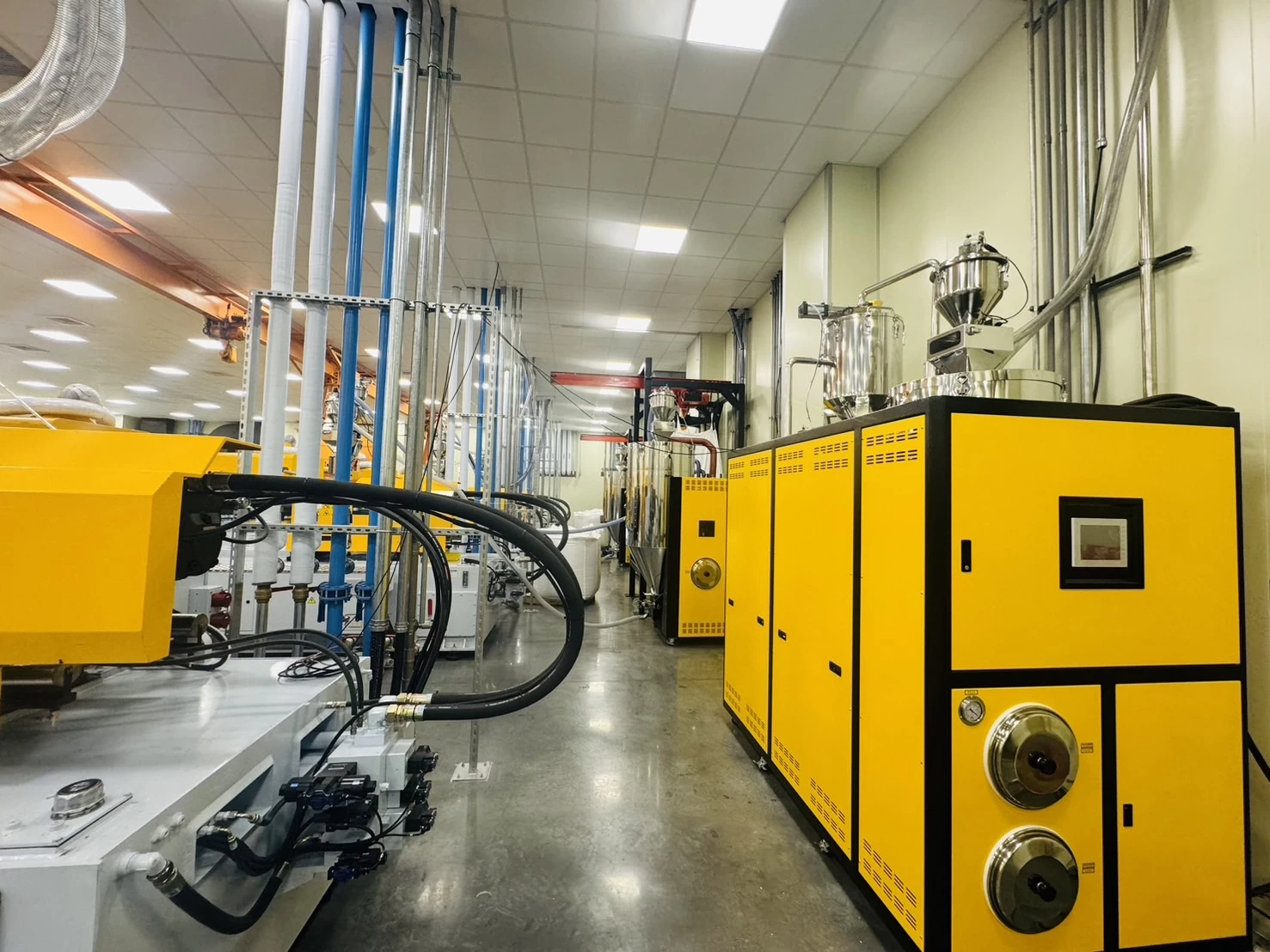 A bright yellow and black Vacuum Dehumidifying Dryer system installed along an industrial factory production line with overhead piping.