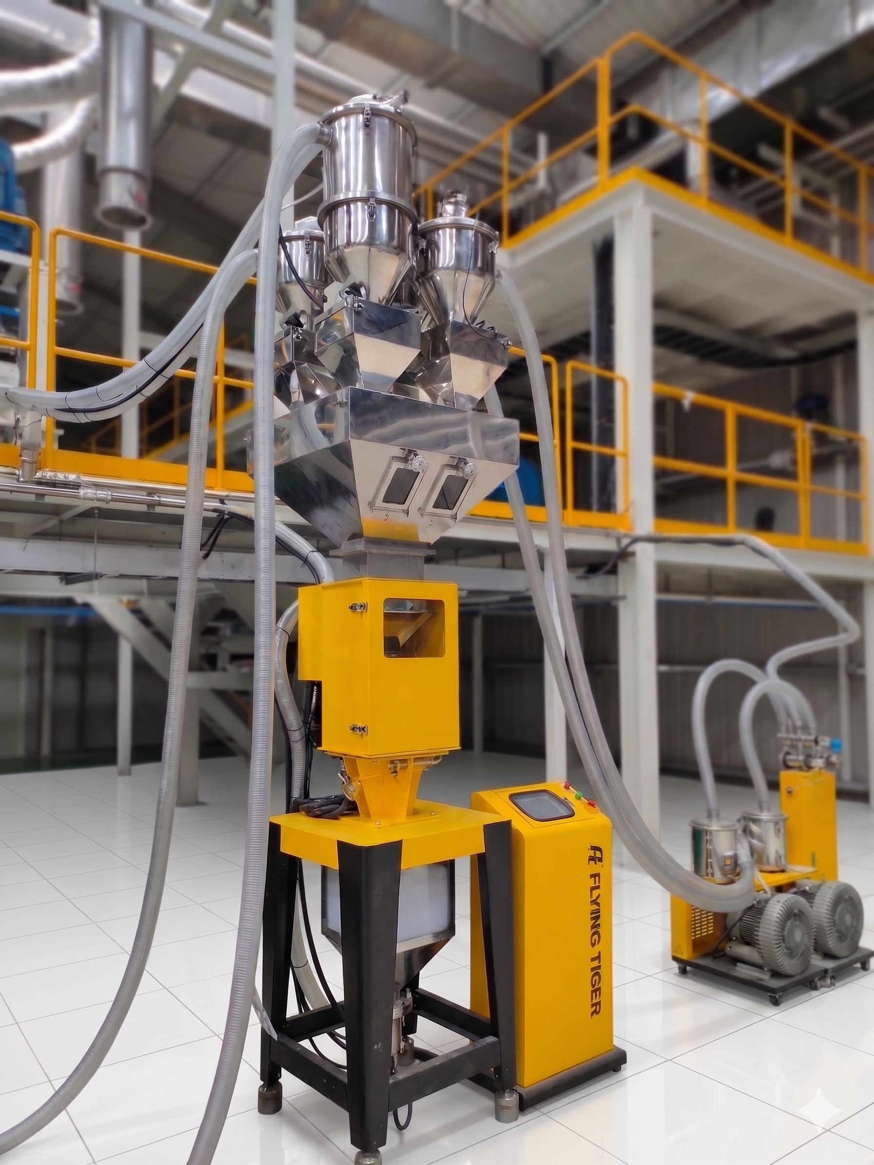 Flying Tiger Gravimetric Dosing Blender system installed on a factory floor. The top section features multiple central material receivers connected to flexible vacuum conveying tubes, feeding into a large metal collection bin. The unit has a yellow touch-screen control panel box.