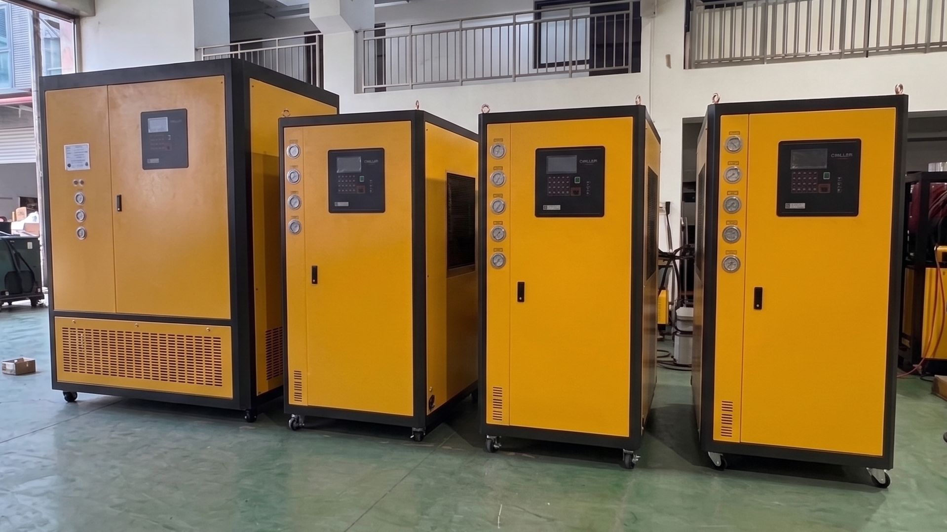 A row of industrial air-cooled chiller units with control panels on a factory floor, waiting for delivery.